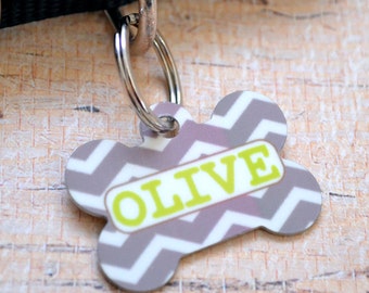 May include: A gray and white chevron patterned bone-shaped dog tag with a green "OLIVE" name tag.