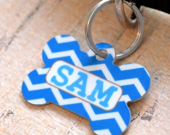 May include: Blue and white chevron dog bone shaped tag with a silver ring and the name "SAM" printed in a white rectangle.
