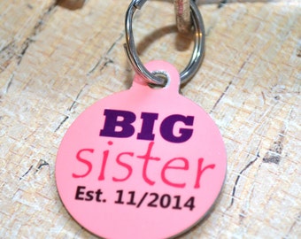 May include: Pink round dog tag with the text "BIG sister Est. 11/2014" in black and pink lettering.
