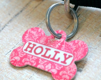 May include: Pink bone-shaped dog tag with a white floral pattern and the name "HOLLY" printed in red.