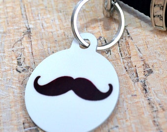 May include: White round dog tag with a black mustache design. The tag is attached to a silver ring.