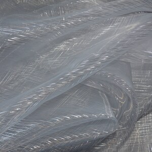 May include: A close-up of a sheer, light blue fabric with a subtle, shimmering quality. The fabric has a delicate, woven texture with silver metallic threads creating a crisscross pattern. The material is lightweight and appears to be slightly translucent.