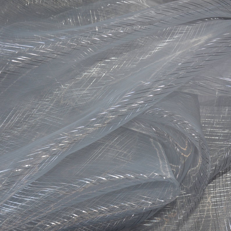 May include: A close-up of a sheer, light blue fabric with a subtle, shimmering quality. The fabric has a delicate, woven texture with silver metallic threads creating a crisscross pattern. The material is lightweight and appears to be slightly translucent.