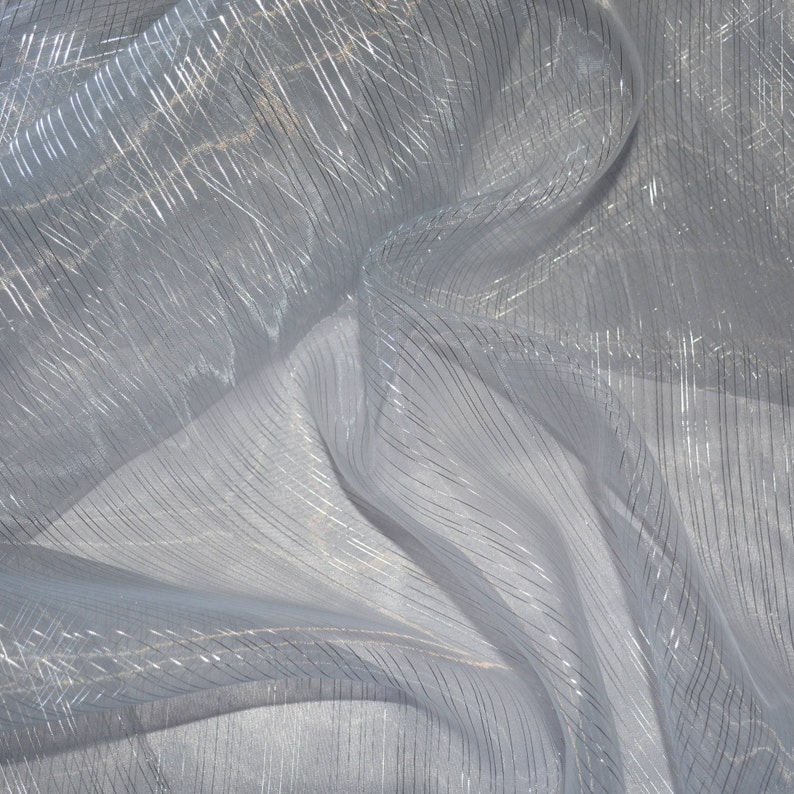 May include: A close-up of a sheer, white fabric with silver metallic stripes. The fabric is draped and folded, creating shadows and highlights. The stripes run both vertically and diagonally across the material, adding texture and visual interest.
