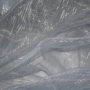 May include: Close-up of a sheer, light blue fabric with silver metallic stripes. The fabric is draped and folded, creating soft shadows and highlights. The stripes run diagonally across the material, adding texture and visual interest. Ideal for apparel or home decor.