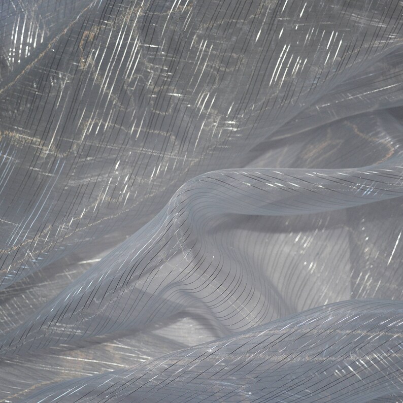 May include: Close-up of a sheer, light blue fabric with silver metallic stripes. The fabric is draped and folded, creating soft shadows and highlights. The stripes run diagonally across the material, adding texture and visual interest. Ideal for apparel or home decor.