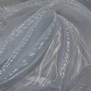 May include: A close-up of a sheer, light gray fabric with a subtle, shimmering quality. The fabric is draped and folded, revealing a pattern of thin, silver metallic stripes that run diagonally across the material. The fabric appears delicate and lightweight.