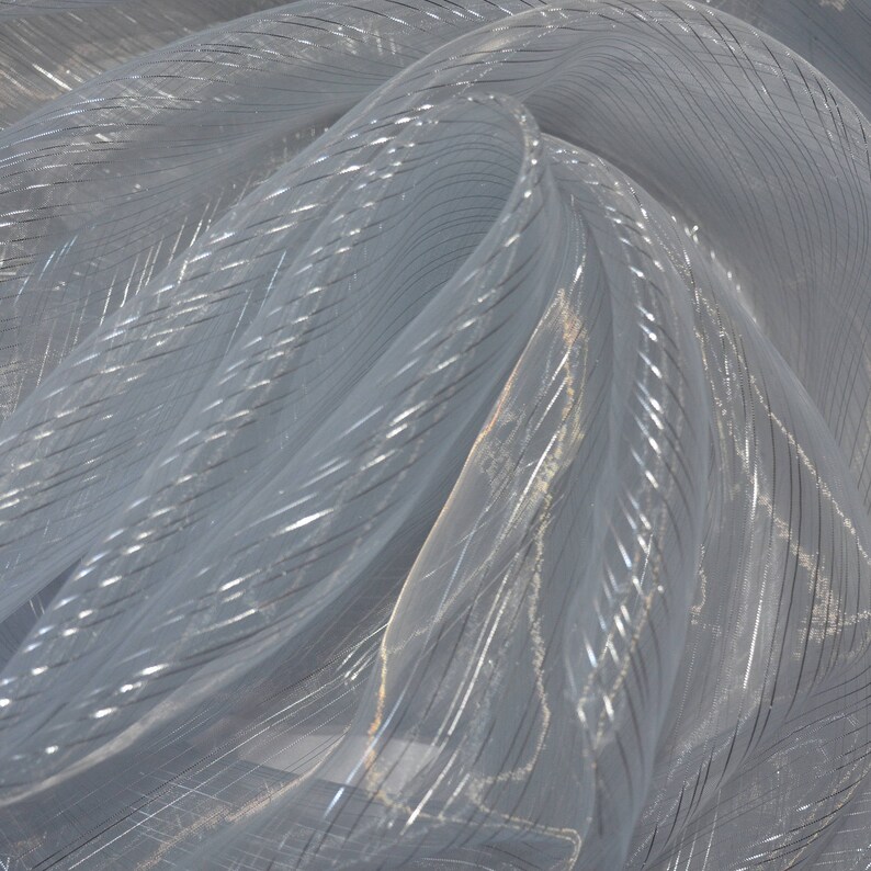 May include: A close-up of a sheer, light gray fabric with a subtle, shimmering quality. The fabric is draped and folded, revealing a pattern of thin, silver metallic stripes that run diagonally across the material. The fabric appears delicate and lightweight.