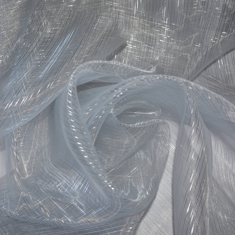 May include: A close-up of a light blue, sheer fabric with a subtle, shimmering effect. The fabric is draped and folded, creating soft curves and shadows. Thin, silver-colored lines run across the material, adding texture and visual interest.