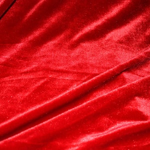 May include: Close-up of a vibrant red velvet fabric. The material is soft and plush, with subtle light reflections highlighting the folds and texture. The rich color and sheen suggest a luxurious textile.