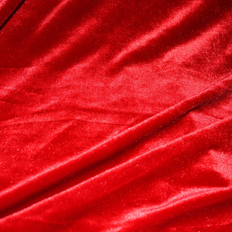 May include: Close-up of a vibrant red velvet fabric. The material is soft and plush, with subtle light reflections highlighting the folds and texture. The rich color and sheen suggest a luxurious textile.