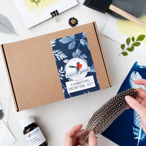May include: A brown cardboard box with a blue and white label that says "Love to print Cyanotype Printing Kit". The box is surrounded by various items, including a feather, a bottle of liquid, and a piece of paper with a blue and white print.