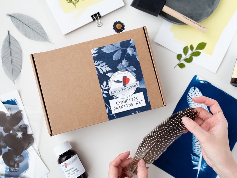 May include: A brown cardboard box with a blue and white label that says "Love to print Cyanotype Printing Kit". The box is surrounded by various items, including a feather, a bottle of liquid, and a piece of paper with a blue and white print.