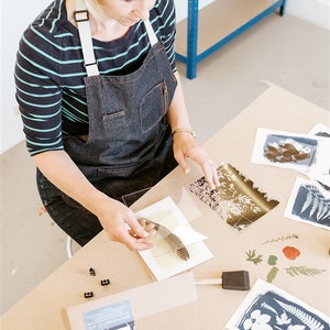 May include: A person is working on a cyanotype art project. They are using a feather to press a leaf onto a piece of paper. There are several other cyanotype prints on the table, as well as a box labeled "Cyanotype Kit".