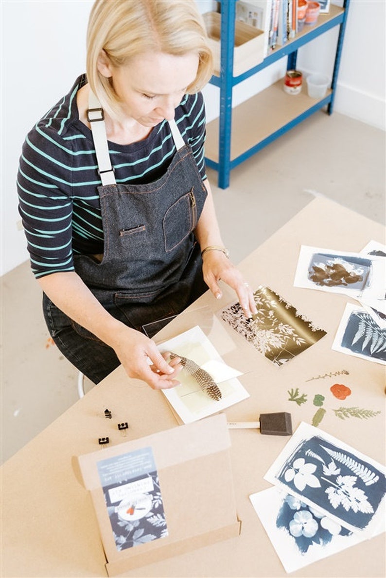 May include: A person is working on a cyanotype art project. They are using a feather to press a leaf onto a piece of paper. There are several other cyanotype prints on the table, as well as a box labeled "Cyanotype Kit".