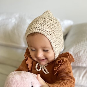 May include: A baby wearing a beige crocheted bonnet with ear flaps and a brown ruffled sweater. The baby is holding a pink plush toy.