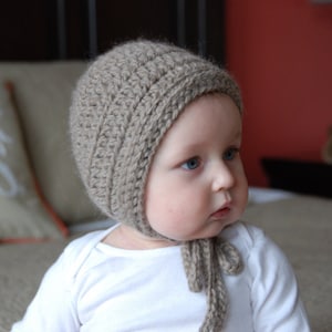 May include: A baby wearing a brown crocheted bonnet with a long tie. The bonnet has a simple stitch pattern and is tied under the chin.