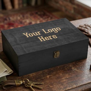 Puede incluir: Una caja de madera negra con el texto "Your Logo Here" grabado en la tapa. La caja tiene un cierre de latón y está sobre una superficie de madera. Un llavero de cuero con llaves está en primer plano.