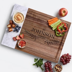 May include: A rectangular wooden serving board with a white marble-patterned side, engraved with the text "the JOHNSON FAMILY" and leaf motifs. The board features a juice groove and is arranged with bowls of hummus, tomatoes, nuts, fruit, and vegetables.