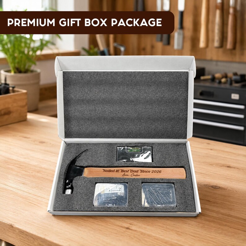 May include: A white gift box containing a claw hammer with a wooden handle engraved with the text Nailed it! Best Dad Since 2026 Love, Caden, a small spirit level, a clear container of nails, and a clear container of screws in foam inserts.