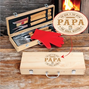 May include: A wooden barbecue tool set with a red heat-resistant glove. The set includes various grilling utensils and is housed in a light-colored wooden case. The case is engraved with the words "PAPA The Grill Master".