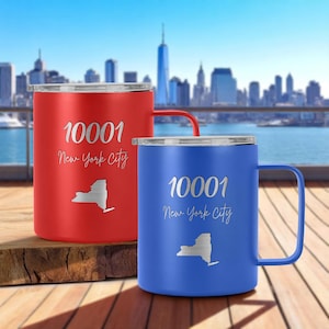 May include: Two insulated travel mugs, one red and one blue, with silver lids and handles. Each mug features the text "10001 New York City" and a New York state outline. The mugs are on a wooden surface with a city skyline in the background.