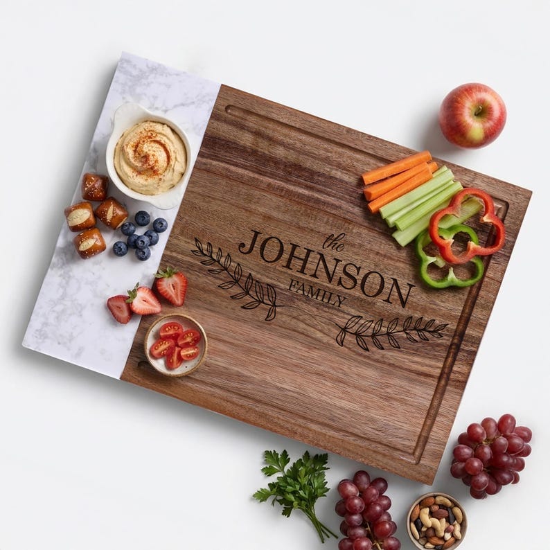 May include: A rectangular wooden serving board with a white marble-patterned side, engraved with the text "the JOHNSON FAMILY" and two leaf branches. The board is arranged with a small bowl of dip, blueberries, strawberries, celery, carrots, and peppers.