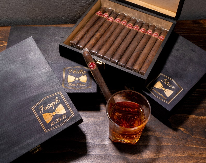 May include: Three black wooden cigar boxes with gold bowtie and text engravings. The boxes are labeled 'Joseph 10.25.21', 'Phillip Best Man', and 'Store Groomswoman'. The top box is open and contains a stack of brown cigars. A glass of amber liquid sits on a wooden surface.