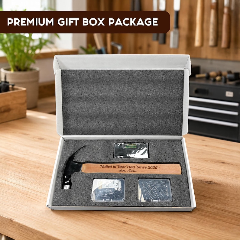 May include: A white gift box containing a claw hammer with a wooden handle engraved with the text Nailed it! Best Dad Since 2026 Love, Caden, a small spirit level, a clear container of nails, and a clear container of screws in foam inserts.