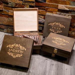May include: Four wooden cigar boxes with engraved designs. The boxes are brown and have gold lettering. The boxes are labeled 'Groom Andrew', 'Groomsman Marc', 'Groomsman Tyler', and 'Groomsman [unreadable name]'. The boxes are arranged in a pyramid shape on a wooden surface.
