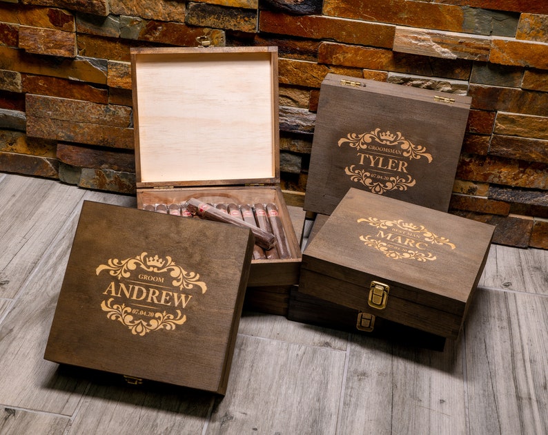 May include: Four wooden cigar boxes with engraved designs. The boxes are brown and have gold lettering. The boxes are labeled 'Groom Andrew', 'Groomsman Marc', 'Groomsman Tyler', and 'Groomsman [unreadable name]'. The boxes are arranged in a pyramid shape on a wooden surface.
