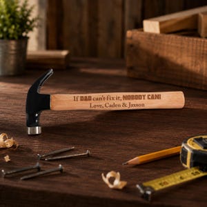 May include: A claw hammer with a black metal head and a light wood handle. The handle is engraved with the text "If DAD can't fix it, NOBODY CAN! Love, Caden & Jaxson". The hammer is positioned horizontally on a dark surface with scattered nails.