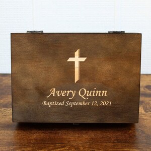 K&ouml;nnte beinhalten: Dunkelbraune Holzkiste mit einem hellfarbigen Kreuz auf der Vorderseite. Der Name "Avery Quinn" und das Datum "Baptized September 12, 2021" sind ebenfalls eingraviert. Die Kiste hat einen Klappdeckel und Metallbeschl&auml;ge.