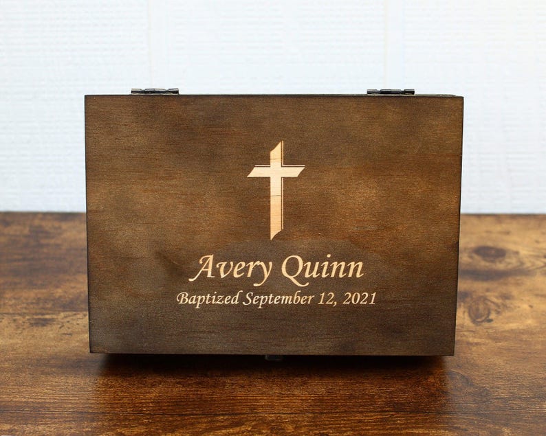 K&ouml;nnte beinhalten: Dunkelbraune Holzkiste mit einem hellfarbigen Kreuz auf der Vorderseite. Der Name "Avery Quinn" und das Datum "Baptized September 12, 2021" sind ebenfalls eingraviert. Die Kiste hat einen Klappdeckel und Metallbeschl&auml;ge.