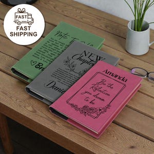 May include: Three personalized journals in green, gray, and pink, with embossed text and floral designs. The journals have the words "Note To", "New Chapter", and "Be the Reflection you dream to be" on the covers. The journals are on a wooden surface.