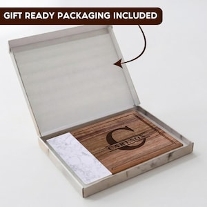 May include: A rectangular wooden cutting board with a white marble-patterned accent on one side, featuring a laser-engraved large letter C and the name CARLSON. The board is presented inside an open white box with a gift ready packaging label.