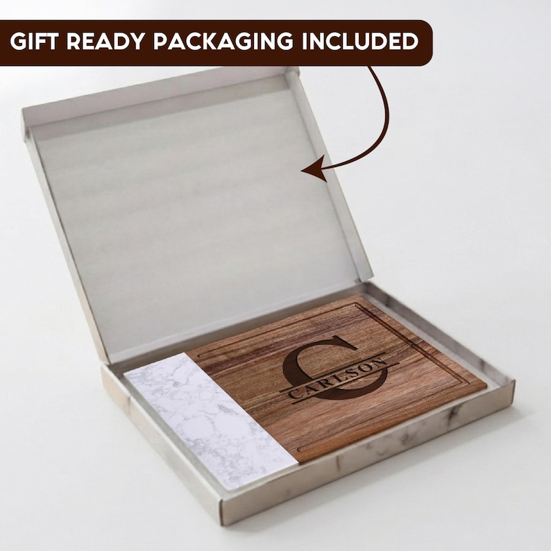 May include: A rectangular wooden cutting board with a white marble-patterned accent on one side, featuring a laser-engraved large letter C and the name CARLSON. The board is presented inside an open white box with a gift ready packaging label.
