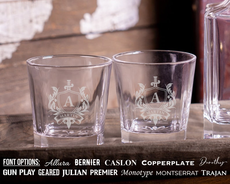 May include: Two clear glass tumblers with a square base. Each tumbler is etched with a crest featuring a sword, shield, and the letter "A" with the name "Martin" below. The text "FONT OPTIONS: Allura BERNIER CASLON COPPERPLATE Dorothy GUN PLAY GEARED JULIAN PREMIER Monotype MONTSERRAT TRAJAN" is displayed below the tumblers.