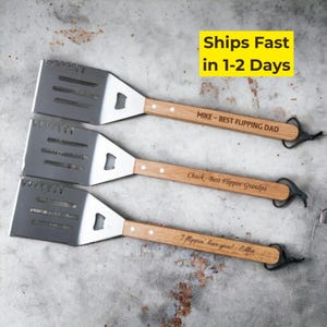 May include: Three stainless steel grilling spatulas with wooden handles. Each spatula has a bottle opener and a loop for hanging. The handles are engraved with phrases like "Best Flippin' Dad" and "I flppin' love you!"
