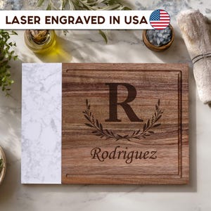 May include: A rectangular cutting board featuring a natural wood grain section and a white marble-patterned side. The wood portion is laser engraved with a large letter R, a decorative leaf wreath, and the name Rodriguez in script font.