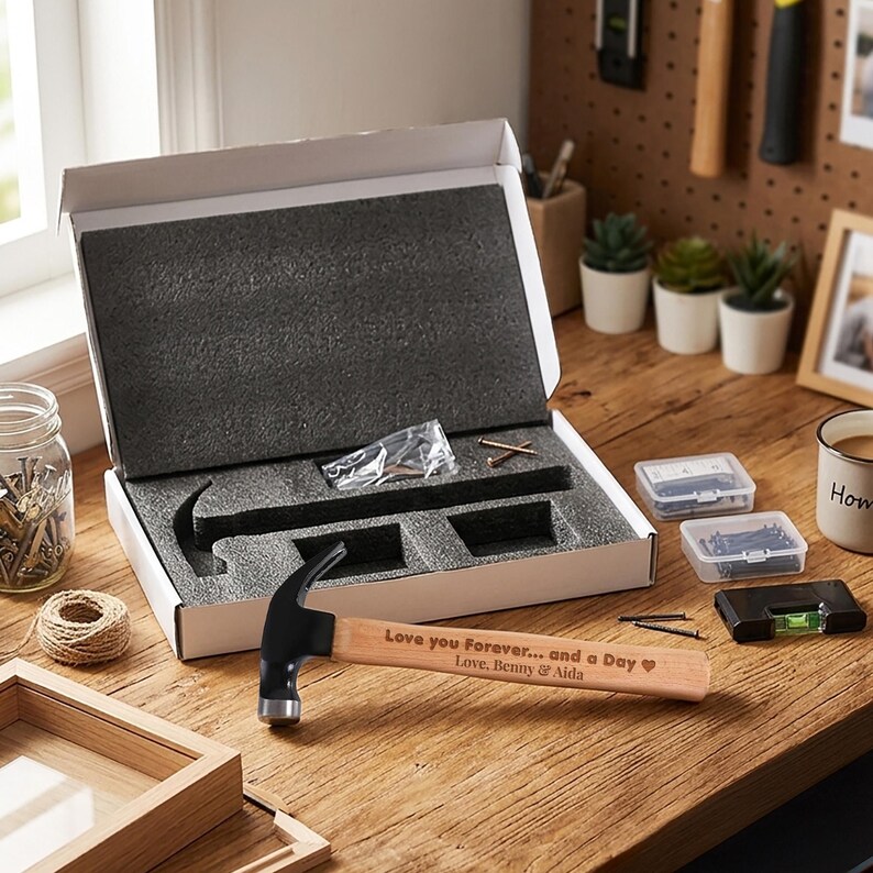 May include: A claw hammer with a light wood handle featuring the engraved text "Love you Forever... and a Day Love, Benny & Aida" with a heart symbol. The hammer is placed in front of an open white cardboard gift box with black foam inserts.