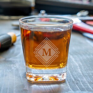 May include: A clear glass rocks glass with a geometric design etched on the side. The design features a diamond shape with the letter "M" in the centre. The glass is filled with a dark amber liquid.