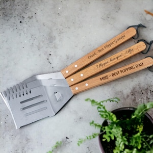 Op de afbeelding: Drie roestvrijstalen barbecuespatels met houten handvatten. Elke spatel heeft een andere gegraveerde boodschap: "Chuck - Best Flippin' Grandpa", "I flippin' love you! - Ellie" en "MIKE - BEST FLIPPING DAD".