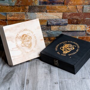 May include: Two wooden boxes with the Golden Steer Steakhouse logo. The logo is a gold steer head with the words 'Golden Steer Steakhouse Las Vegas Est. 1958' around it. One box is light wood and the other is black. The boxes are sitting on a wood floor.