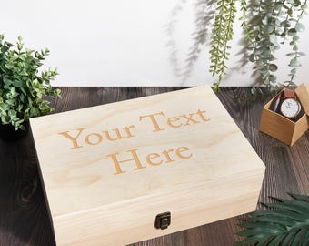 Può includere: Una scatola di legno color chiaro con il testo inciso "Your Text Here" sul coperchio. La scatola ha una chiusura in metallo ed è appoggiata su una superficie di legno scuro. Un orologio in una scatola di legno è visibile sullo sfondo.