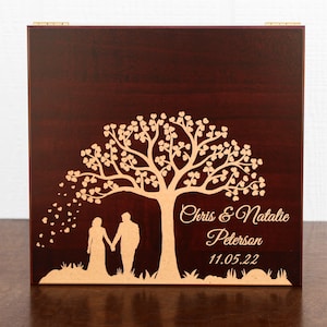 May include: A dark brown wooden box with a gold engraved design of a tree with hearts, two people holding hands, and the text "Chris & Natalie Peterson 11.05.22".