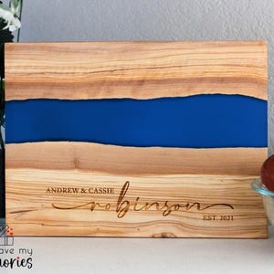May include: Wooden cutting board with a blue resin river design. The board is engraved with the names "Andrew & Cassie" and the last name "Robinson", with "EST. 2021" below. The board is made of light brown wood.