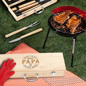 May include: A wooden barbecue tool set and a small red and black grill with ribs cooking over flames. The set includes a fork, knife, and tongs with wooden handles. The wooden box is engraved with "PAPA The Grill Master".
