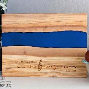 K&ouml;nnte beinhalten: Holzschneidebrett mit einem blauen Harzfluss-Design. Das Brett ist mit den Namen "Andrew & Cassie Robinson" und "EST. 2021" graviert. Das Brett besteht aus hellbraunem Holz mit einer dunkelblauen Mitte. Die Worte "engrave my memories" stehen unten links.