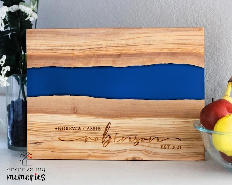 K&ouml;nnte beinhalten: Holzschneidebrett mit einem blauen Harzfluss-Design. Das Brett ist mit den Namen "Andrew & Cassie Robinson" und "EST. 2021" graviert. Das Brett besteht aus hellbraunem Holz mit einer dunkelblauen Mitte. Die Worte "engrave my memories" stehen unten links.